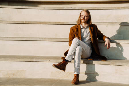 White ginger man wearing jacket and eyeglasses sitting on stairs outdoorsの写真素材
