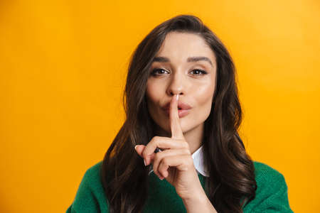 Portrait of a smiling casual woman standing over yellow background showing finger over lips