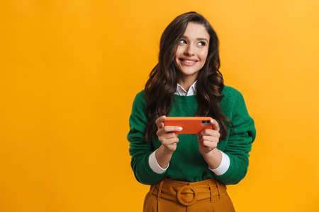 Smiling casual young brunette woman holding mobile phone standing over yellow background playing gamesの写真素材