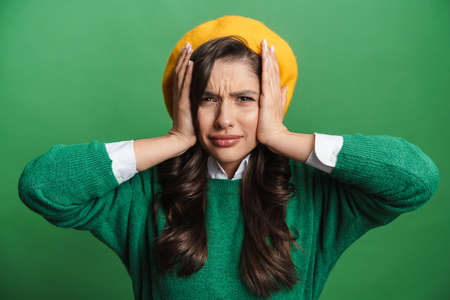 Young brunette woman holding her head while frowning isolated over green background