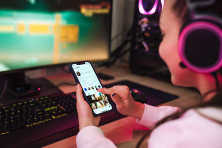 Young happy woman gamer chatting on smartphone while sitting at the desk with computer in dark room...