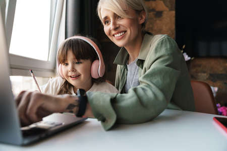 Happy mother helping her daughter with school homework, using laptop computer, study during...