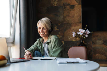 Smiling middle aged blonde woman with short hair studying or working on a video call via laptop computer while sitting at the table at homeの写真素材