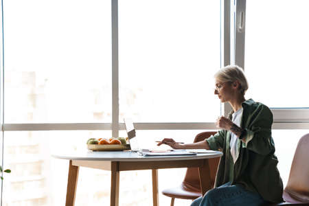 Middle aged blonde woman with short hair studying or working on a video call via laptop computer while sitting at the table at homeの写真素材
