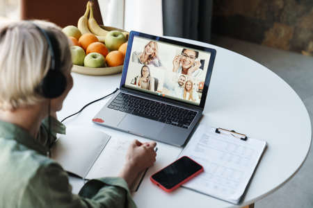 Back view of a smiling mid aged blonde woman on a video call with colleagues via laptop computer...