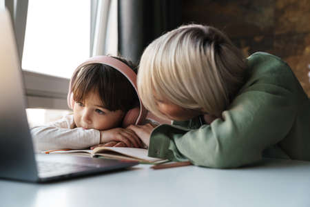 Mother helping her daughter with school homework, using laptop computer, study during quarantine, little girl listening to teacher in headphonesの写真素材