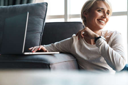 Smiling mid aged blonde woman studying online via laptop computer in the living room
