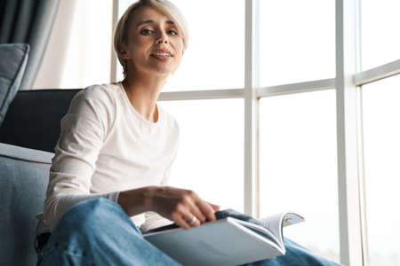 Smiling mid aged blonde woman studying reading book while sitting on a floor in the living room