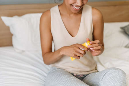 Young black woman smiling and applying hand cream while sitting on bed at home