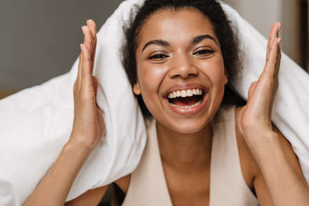 Happy young african woman laying hiding under a blanket on a bed at home laughing