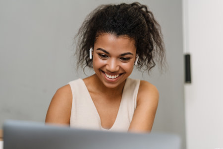 Happy young african woman sitting in bed in the bedroom wearing earphones talking on a video call...