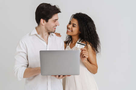 Multiracial smiling couple posing with laptop and credit card isolated over white background