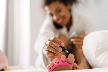 Smiling young african woman in bathrobe sitting on a bed painting her nails in the bedroom talking on mobile phoneの写真素材
