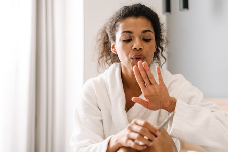 Smiling young african woman in bathrobe sitting on a bed filing her nails in the bedroomの写真素材