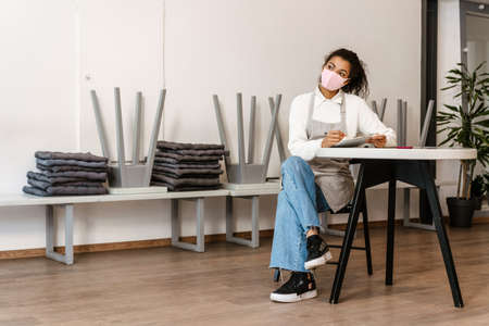 Young black waitress wearing face mask writing down notes on clipboard on workplaceの写真素材