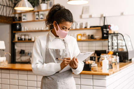 Young black waitress wearing face mask writing down notes on clipboard in cafeの写真素材