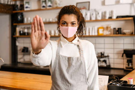 Young black waitress in face mask showing stop gesture by cafe counter indoorsの写真素材