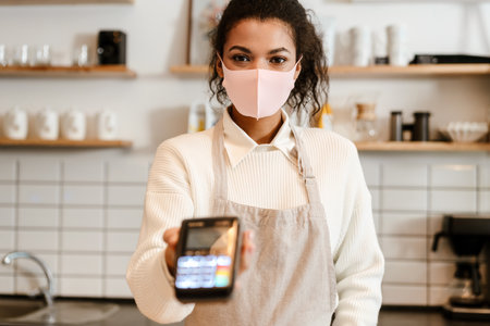 Young black waitress in face mask working with payment terminal at cafe counter indoors