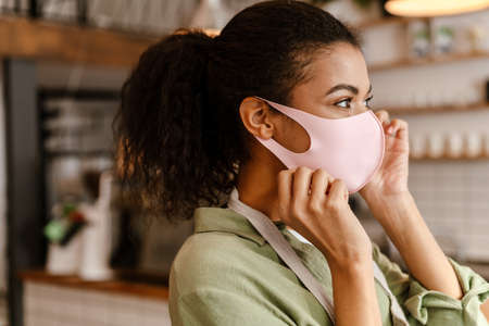 Black waitress wearing face mask while getting ready for work at cafe indoorsの写真素材
