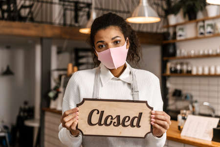 Young black waitress in face mask showing close sign board at cafe indoors
