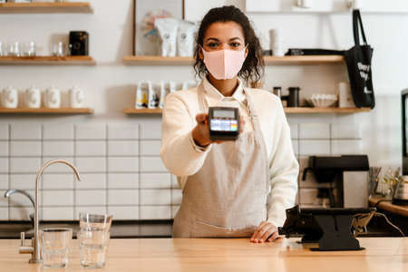 Young black waitress in face mask working with payment terminal at cafe counter indoorsの写真素材