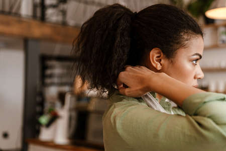 Young black waitress wearing apron while getting ready for work in cafe indoors