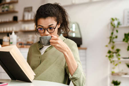 Young black woman in eyeglasses reading book while drinking coffee at cafe indoors