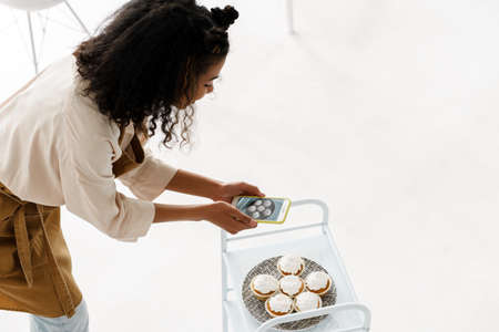 Black young woman in apron taking photo of her muffins at home