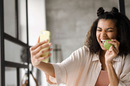 Young happy african american woman holding smart phone making a vlog from home biting green apple