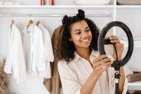 Black young woman smiling while taking selfie footage on cellphone at wardrobeの写真素材