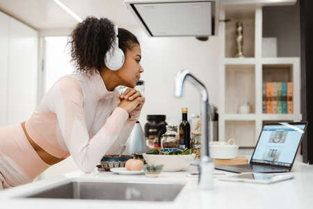 Black woman in headphones using laptop while making smoothie at home