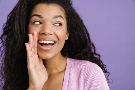 Close up of a happy young african woman in casual clothes standing over violet background, looking awayの写真素材