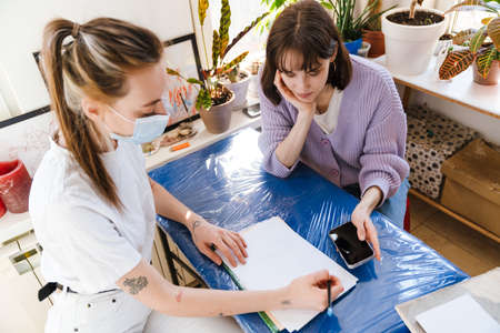 Young white women using cellphone while doing sketches in workshop