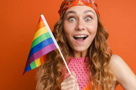 Young ginger woman expressing surprise while posing with rainbow flag isolated over orange wall