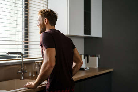 Attractive happy young man looking in the window while standing at the kitchen sinkの写真素材