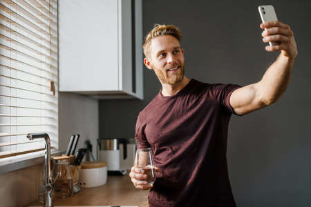 Attractive happy young man taking a selfie while drinking water from a glass at homeの写真素材