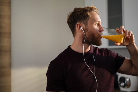 Attractive healthy young man having breakfast while standing in the kitchen, drinking juice, listening to music with earphonesの写真素材