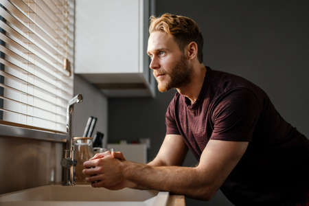 Attractive young man looking in the window while standing at the kitchen sinkの写真素材