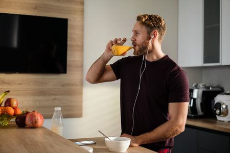Attractive healthy young man having tasty breakfast while standing in the kitchen, drinking juice, listening to music with earphonesの写真素材