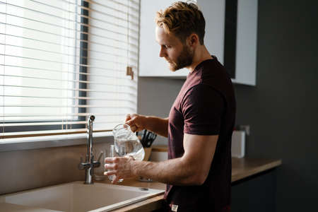 Attractive young man pouring water from a juf in a glass while standing at the kitchen sinkの写真素材