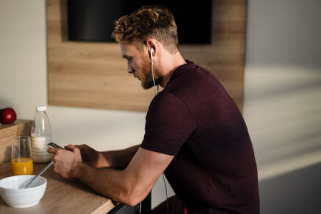 Handsome young man listening to music with earphones, browsing on mobile phone while sitting in the kitchenの写真素材