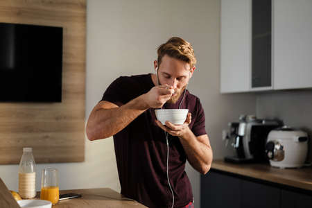 Attractive healthy young man having tasty breakfast while standing in the kitchen, holding bowl, listening to music with earphonesの写真素材