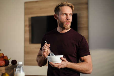 Attractive healthy young man having tasty breakfast while standing in the kitchen, holding bowlの写真素材