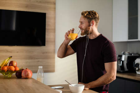 Attractive healthy young man having tasty breakfast while standing in the kitchen, drinking juice, listening to music with earphonesの写真素材