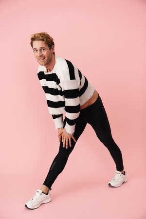 Ginger young man wearing tights smiling while doing exercise isolated over pink wall