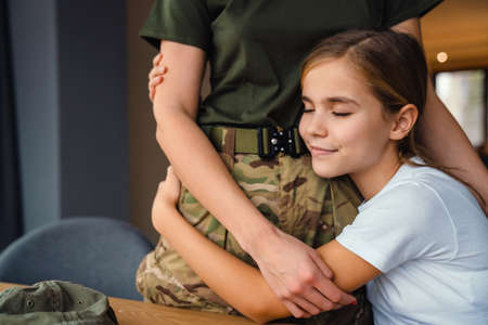 Beautiful military woman hugging with her smiling daughter at homeの写真素材