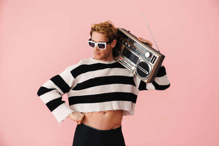 Ginger young man wearing sunglasses posing with retro tape recorder isolated over pink wall