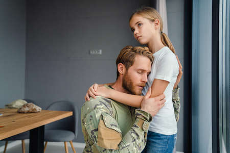 Masculine military man hugging her daughter while kneeling indoorsの写真素材