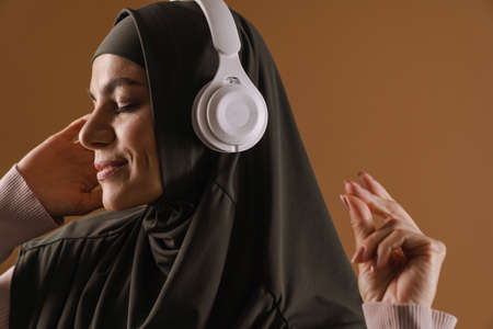 Young muslim woman in hijab listening music with headphones isolated over brown background