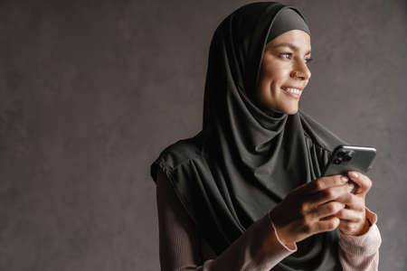 Young muslim woman in hijab smiling while using mobile phone isolated over grey backgroundの写真素材